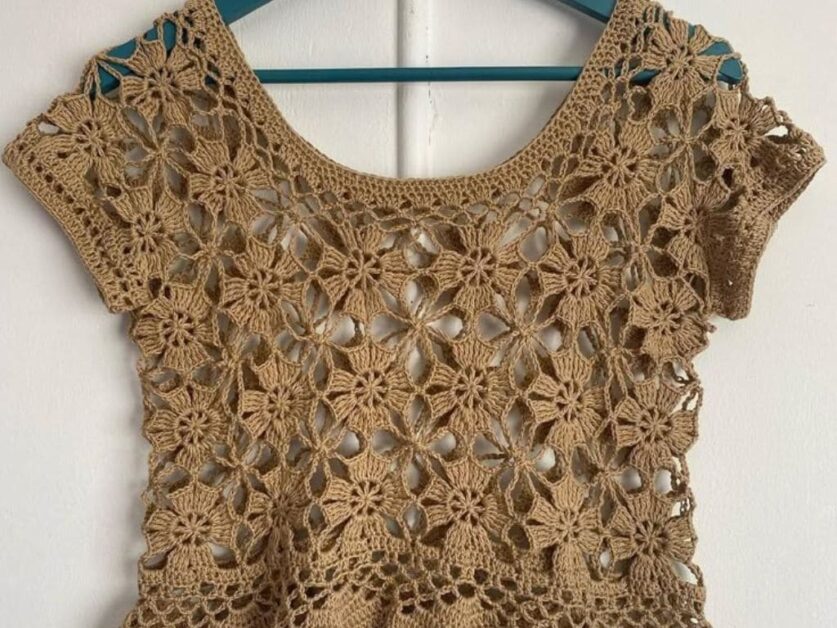 Blusa de crochê marrom clara com padrão floral pendurada em um cabide azul.