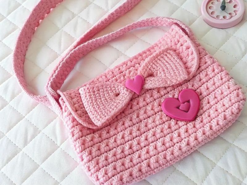 Bolsinha de crochê infantil rosa com alça e detalhes de laço e corações.