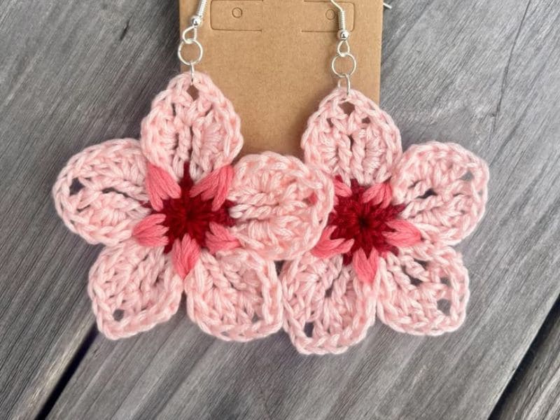Par de brincos de crochê em formato de flor rosa com centro vermelho, pendurados em suportes de metal prateado.