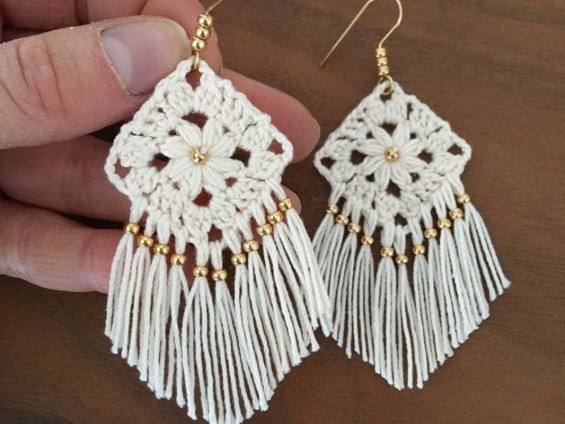 Close-up de um par de brincos de crochê brancos e elegantes sendo segurados, com detalhes em dourado.