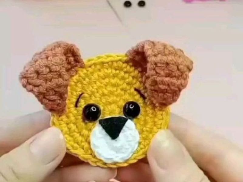 Aplique de crochê com a forma da cabeça de um cachorrinho amarelo com orelhas marrons, olhos pretos e nariz triangular preto, sendo segurado por duas mãos.