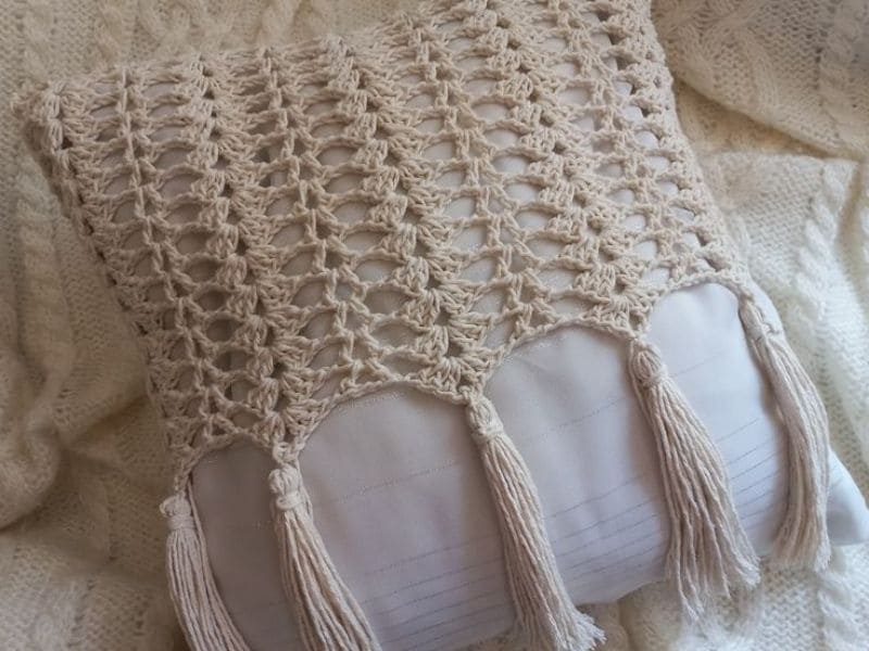 Capa de almofada de crochê moderna, bege claro, com detalhes em franja e padrão vazado sobre fundo branco.