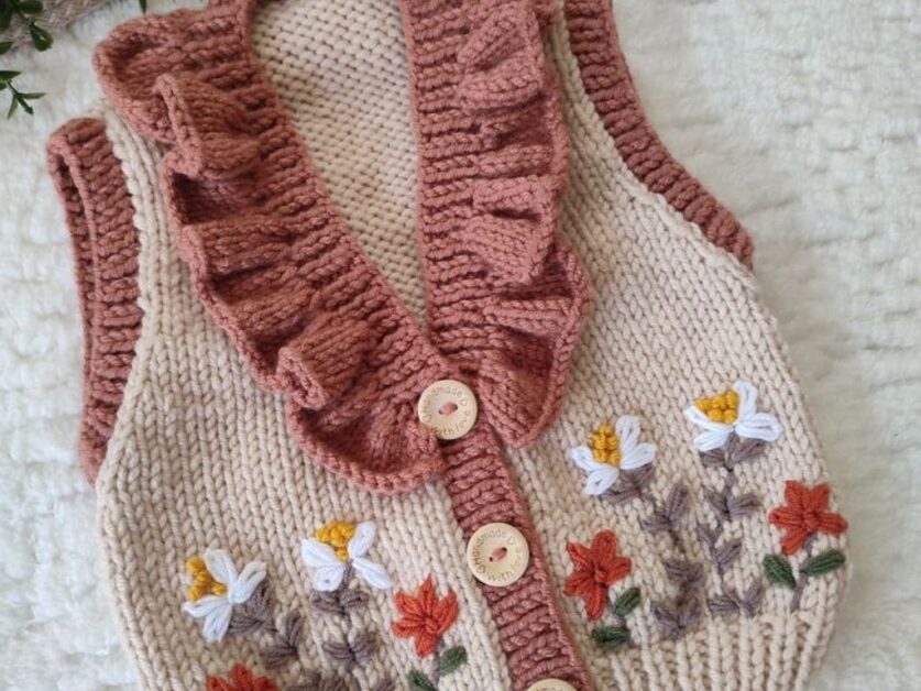 Casaquinho de bebê em crochê bege com detalhes em crochê rosa e bordado floral, fechado por botões de madeira.