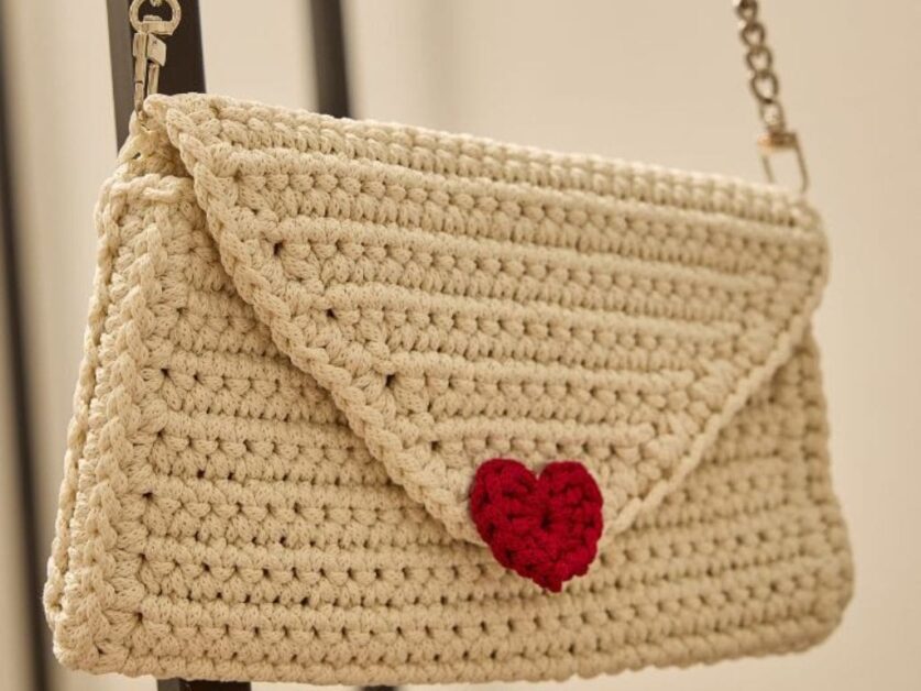 Bolsa de crochê bege em formato de envelope com um coração vermelho decorativo.