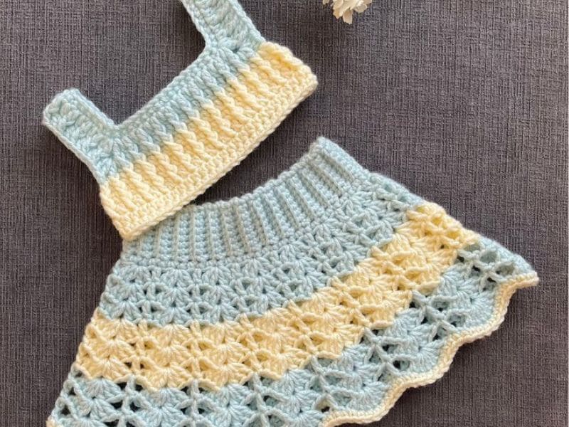 Conjuntinho infantil de crochê azul e amarelo, composto por top e saia, em fundo cinza.