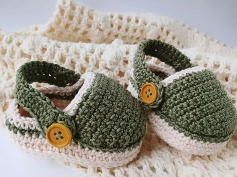 Close-up de um par de sapatinhos de crochê para bebê, em tons de verde e bege, com botões decorativos.
