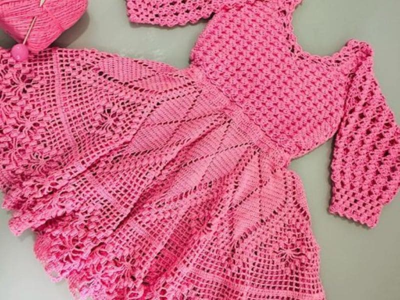 Vestido de crochê infantil rosa com mangas compridas e detalhe floral na saia.