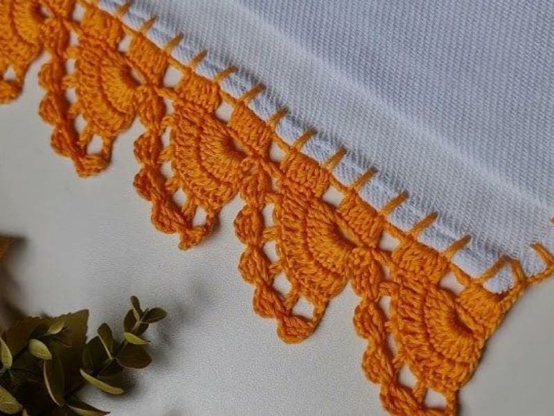Detalhe de um bico de crochê laranja vibrante adornando a borda de um tecido branco, com foco na delicadeza dos pontos.