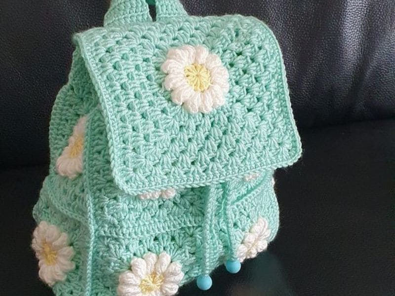 Mochila infantil azul de crochê com detalhes de flores brancas.