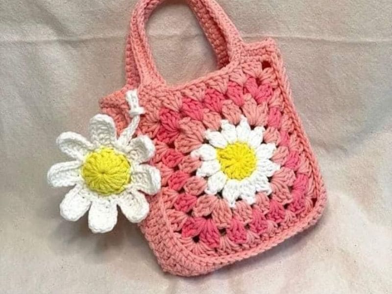 Bolsa de crochê infantil rosa com aplicação de flor de crochê branca e amarela.