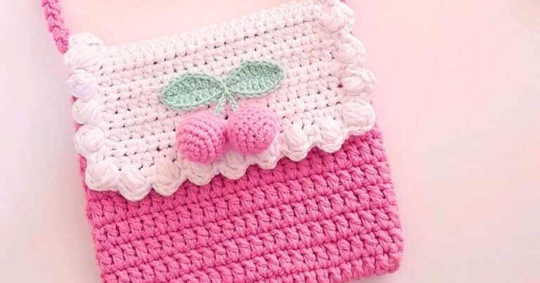 Bolsa infantil de crochê rosa e branca, decorada com cerejas, sobre um fundo rosa.