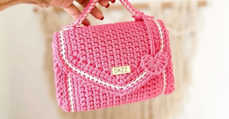 Bolsa de crochê rosa com detalhes brancos e uma etiqueta dourada com a palavra 'DAZZ'.