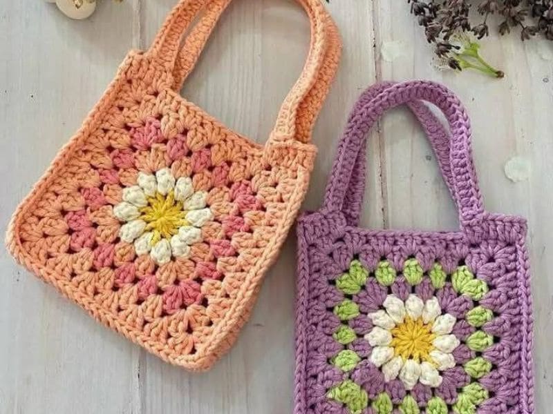 Duas bolsas de crochê infantil coloridas, uma em tons de pêssego e outra lilás, ambas com desenho de flor.