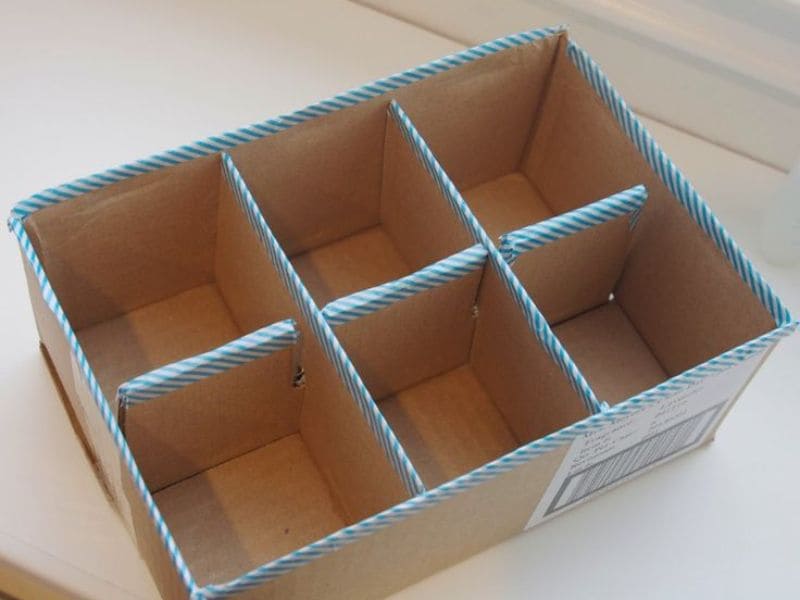 Caixa organizadora de papelão marrom com seis divisórias, decorada com fita adesiva azul e branca.