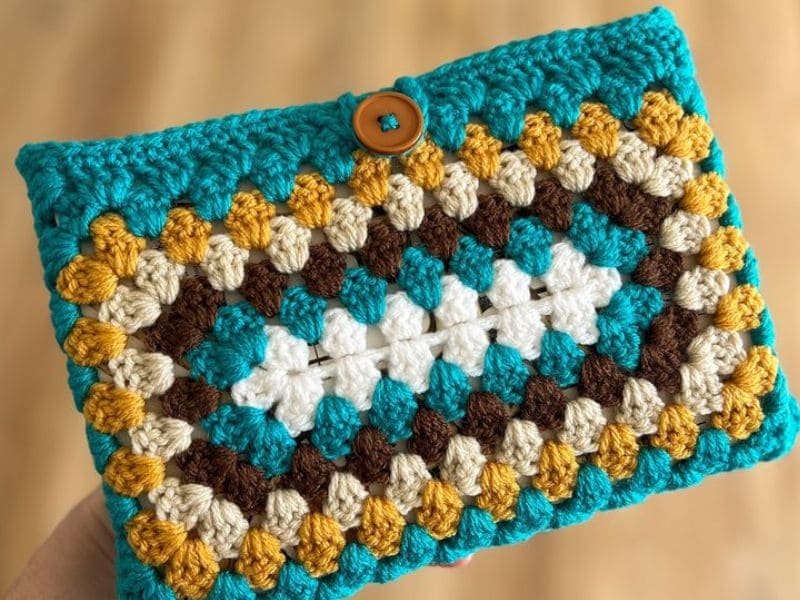 Carteira de crochê com granny squares coloridos em tons de azul, marrom, branco e amarelo, fechada com um botão de madeira.
