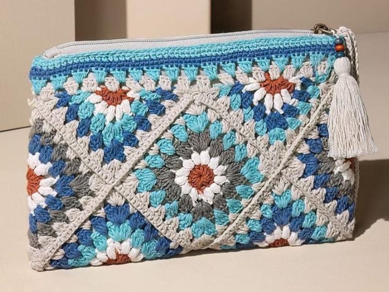 Carteira de crochê com granny squares em tons de azul, branco, cinza e laranja, com zíper e tassel decorativo.
