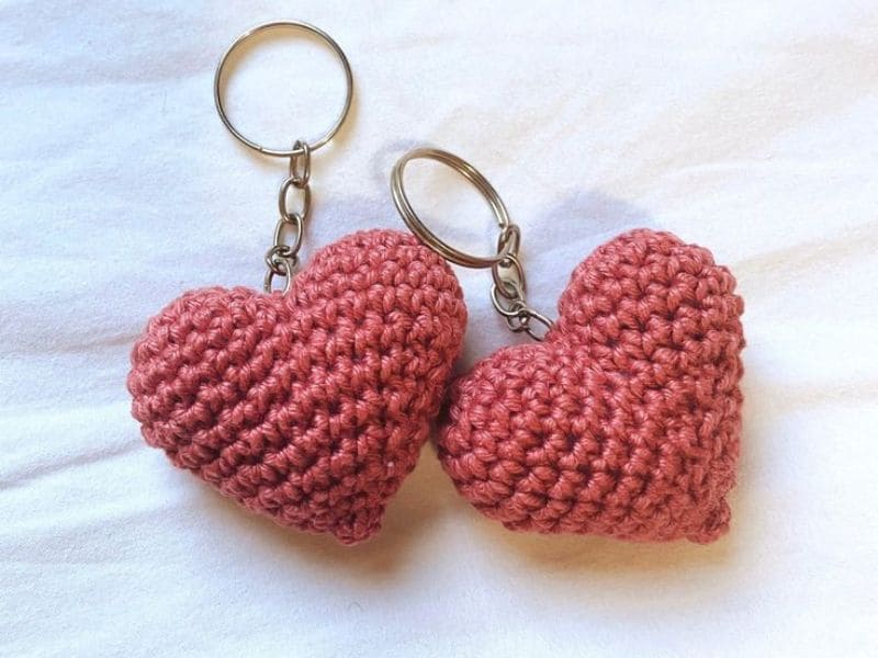 Dois chaveiros de crochê em forma de coração rosa sobre um fundo branco texturizado.