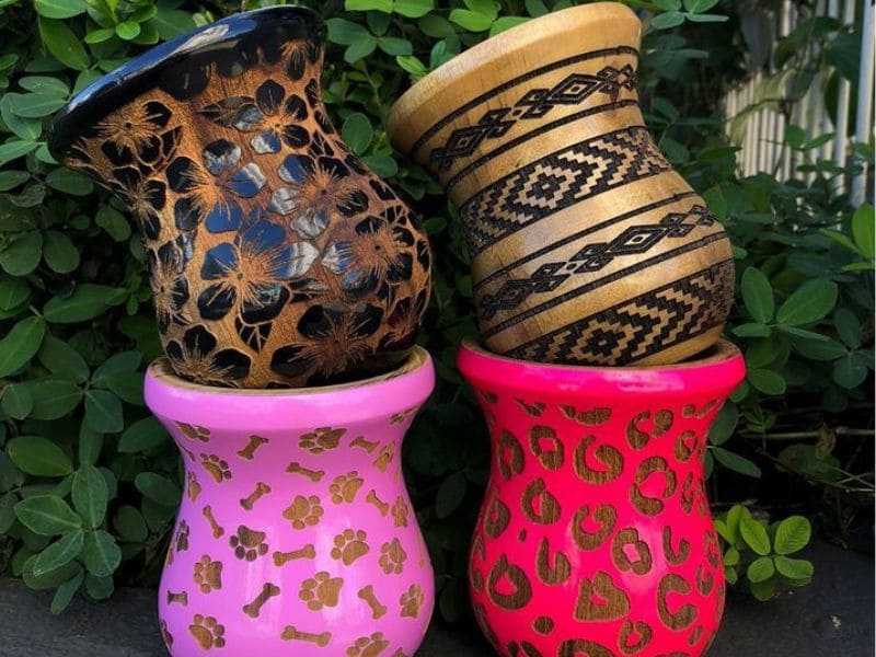 Quatro cuias artesanais decoradas em diferentes estilos, empilhadas em pares sobre um fundo de folhagem verde.