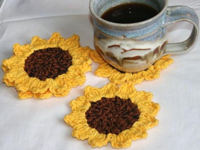 Três descansos de copos em formato de girassol feitos de crochê, um deles com uma xícara de café em cima.