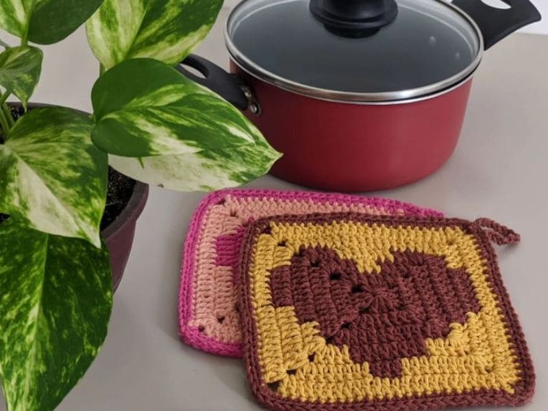 Dois descansos de panela de crochê coloridos com design de coração, panela vermelha com tampa e planta verde ao lado.