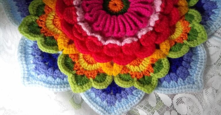Detalhe de uma flor de crochê grande e colorida, com camadas em tons de rosa, vermelho, laranja, amarelo, verde e azul, sobre uma superfície de renda branca.