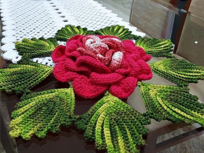 Centro de mesa com grande flor de crochê vermelha e detalhes em crochê verde imitando folhas, sobre uma mesa de vidro com toalha branca.
