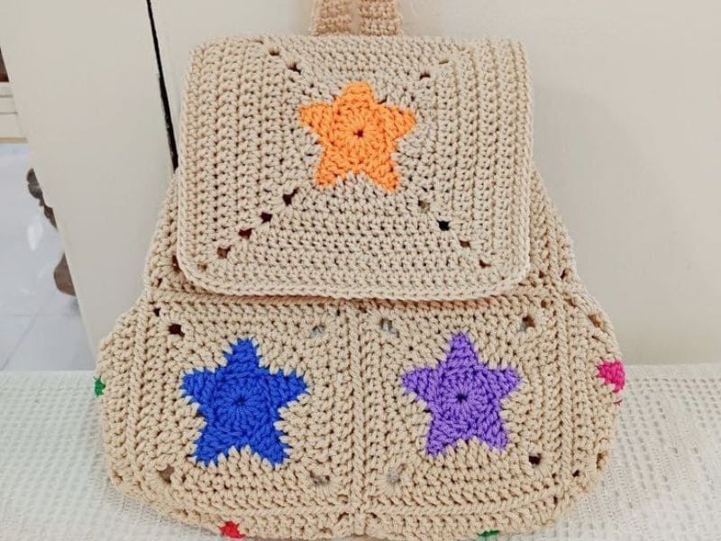 Bolsa infantil de crochê bege em formato de mochila com estrelas coloridas.