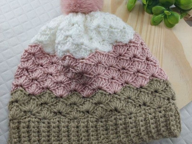 Close-up de uma linda touca de crochê feminina com pompom, em tons de branco, rosa e marrom.