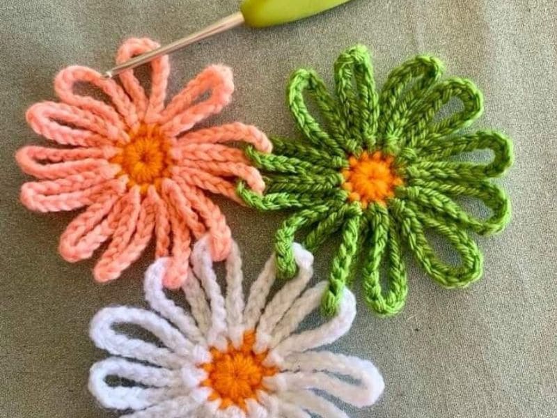 Três lindas flores de crochê em tons de pêssego, verde e branco com centros laranjas, dispostas sobre um fundo texturizado verde, com uma agulha de crochê verde presente.