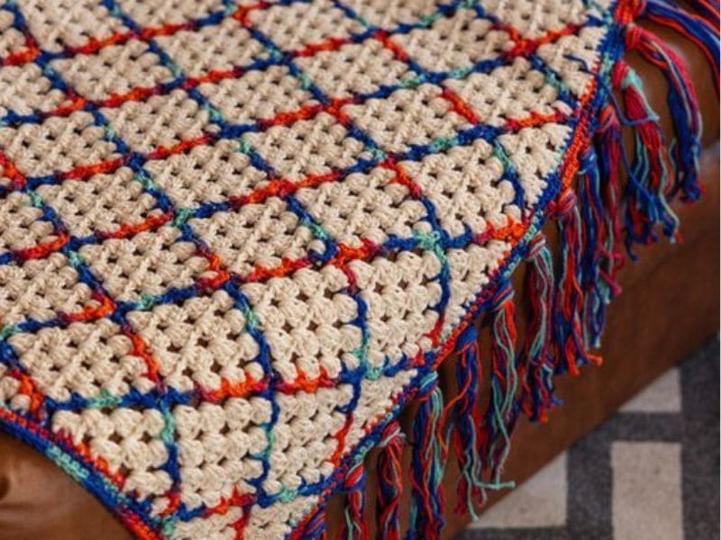 Detalhe de uma manta de crochê artesanal com franjas coloridas, exibindo um padrão geométrico quadriculado.