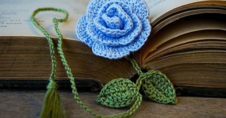 Um livro aberto com um marcador de página de crochê em forma de rosa azul e folhas verdes.