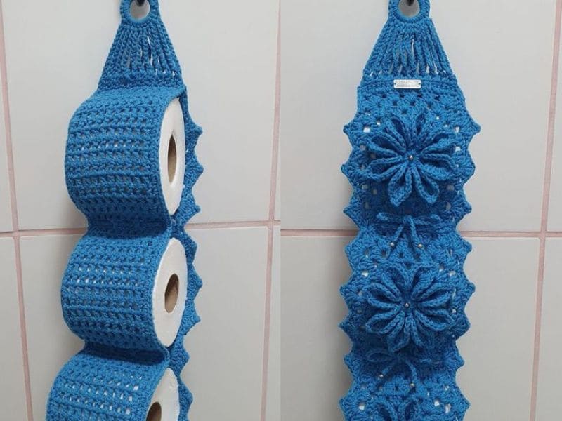 Porta papel higiênico de crochê azul pendurado na parede com três rolos de papel.