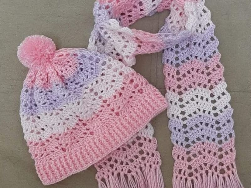 Conjunto de crochê feminino rosa, branco e lilás, composto por uma touca com pompom e um cachecol.