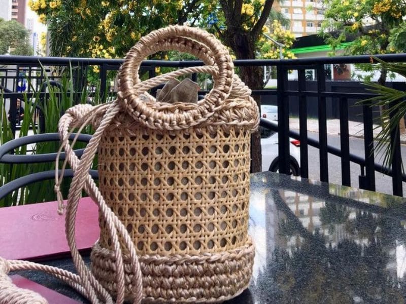 Bolsa de palha artesanal com alça redonda e cordão, sobre uma mesa escura com fundo de vegetação.